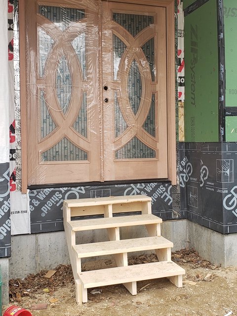Pair of wood entry doors with decorative glass set into a wall that is still wrapped in sheathing, with temporary wooden steps