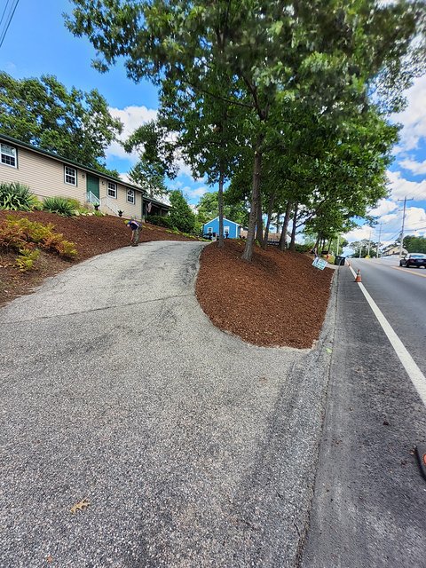 Driveway curving up to a tan house, with freshly spread dark mulch on the slopes beside it and a row of mature trees along the road