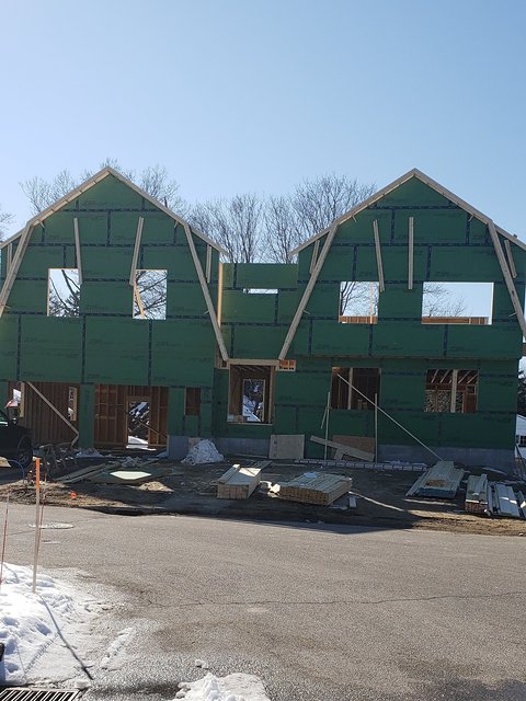 Two-story house under construction with green taped sheathing on the walls, framed gables and window openings, against a clear blue sky